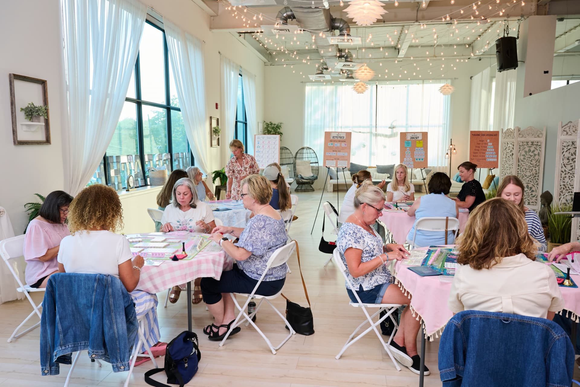 A bright studio filled with tables of students playing mahjong at a Mahj with Sue class
